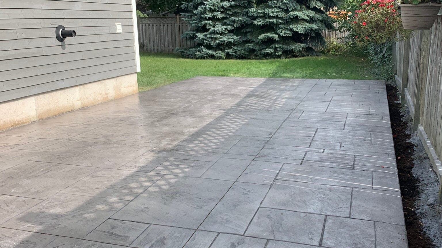 Grey stamped concrete patio in a slate tile pattern running along the side of a house, with a lawn, evergreens and a wooden fence behind it